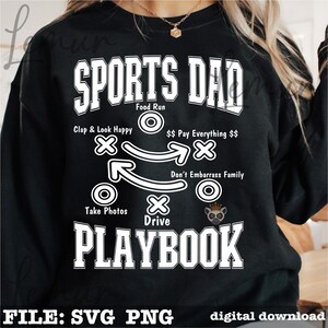 以下が含まれることがあります： 黒いスウェットシャツに、白い文字で「SPORTS DAD PLAYBOOK」と書かれています。デザインには、「Clap & Look Happy」や「Take Photos」などのフレーズを含むフットボールのプレー図が含まれています。