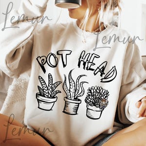 May include: Cream-colored sweatshirt with the text "POT HEAD" above three potted plants. The plants are illustrated in black and white, with a variety of leaf shapes and textures. The sweatshirt is a casual, comfortable style.