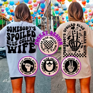 May include: Two white t-shirts with black text and graphics. The front of the shirt says "Somebody's Spoiled Blue Collar Wife". The back of the shirt says "Somebody's Spoiled Blue Collar Wife" with a graphic of a skeleton hand giving the peace sign. The pocket design on the shirt is a checkered heart with dripping paint.