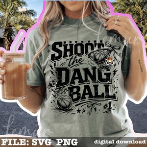 Puede incluir: Camiseta verde claro con el texto negro "SHOOT the DANG BALL". El dise&ntilde;o incluye una pelota de baloncesto, una red y un silbato. La camiseta tambi&eacute;n tiene el n&uacute;mero 1 y estrellas. Un vaso con pajita y bebida se sostiene en la mano izquierda.