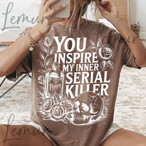 May include: Brown t-shirt with the text "YOU INSPIRE MY INNER SERIAL KILLER" in white. The graphic includes a poison bottle, roses, and a skull with an owl. The shirt has a crew neck and short sleeves.