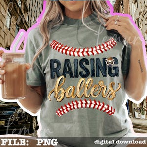May include: A gray t-shirt with the words "RAISING ballers" in black and gold lettering, with baseball stitching above and below the text. The shirt has a relaxed fit.