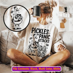 May include: A cream-coloured sweatshirt featuring the text "Pickles and Panic Attacks" and a black and white graphic of a goose holding a jar of pickles. The goose wears a hat and boots. Additional pickle jars are also shown.