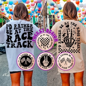 May include: Two white t-shirts with a black graphic print that says "I'd rather be at the race track". One shirt has a checkered heart with dripping paint and the other has a skeleton hand with a peace sign. Both shirts have a smaller circle graphic on the bottom left corner.
