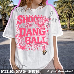 Puede incluir: Camiseta blanca con un dise&ntilde;o rosa de baloncesto. El dise&ntilde;o incluye el texto "SHOOT THE DANG BALL" con una pelota de baloncesto, una red y otros elementos deportivos. La camiseta tambi&eacute;n presenta una peque&ntilde;a ilustraci&oacute;n de un l&eacute;mur coronado.