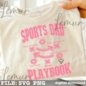 以下が含まれることがあります： クリーム色のTシャツ。ピンク色の文字で「SPORTS DAD PLAYBOOK」と書かれています。デザインには、「Food Run」、「Clap & Look Happy」、「Don't Embarrass Family」などのフレーズを含むフットボールのプレー図が含まれています。キツネザルのイラストも含まれています。