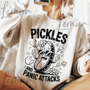 May include: A cream-coloured sweatshirt featuring a black graphic with the text "PICKLES AND PANIC ATTACKS." The design includes a cartoon pickle in sunglasses, peace signs, flowers, stars, and lightning bolts, creating a fun, quirky aesthetic.