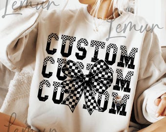 May include: A cream-coloured sweatshirt featuring the word "CUSTOM" in large, black, chequered letters. A large chequered bow is centered below the word. The sweatshirt is a casual, comfortable style.