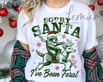 Może przedstawiać: Biała koszulka z zieloną grafiką szopa w czapce Świętego Mikołaja jadącego na reniferze, z napisem "SORRY SANTA I've Been Feral". Czerwone płatki śniegu rozrzucone wokół wzoru. Na dole koszulki napis "FILE: SVG PNG digital download".
