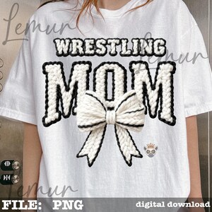 May include: White t-shirt with the words "WRESTLING MOM" in a black and white knitted design. A large white bow with black trim is centered below the word "MOM". A small lemur graphic is on the right side of the shirt.