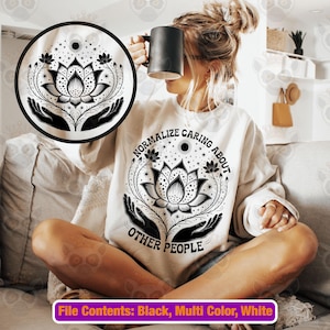 Puede incluir: Una sudadera crema con un diseño de flor de loto en blanco y negro y el texto "Normalize Caring About Other People". El diseño incluye manos, estrellas y un sol. Una taza negra se sostiene en la mano de la persona.
