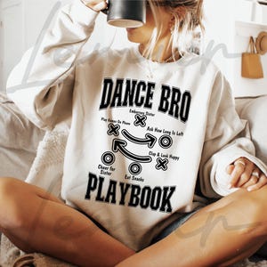 May include: A white sweatshirt with a black graphic that says "Dance Bro" and includes a football playbook with instructions like "Embarrass Sister", "Play Games On Phone", "Ask How Long Is Left", "Cheer for Sister", "Eat Snacks", and "Clap & Look Happy".