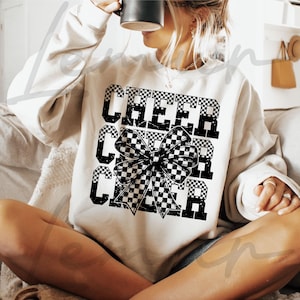 Può includere: Una felpa bianca con un fiocco a scacchi bianco e nero e il testo "CHEER" ripetuto tre volte.