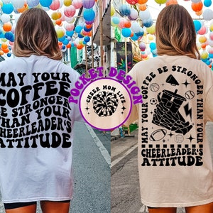 May include: Two white and beige t-shirts with black text and graphics. The front of the white t-shirt says "May your coffee be stronger than your cheerleader's attitude". The back of the beige t-shirt says "May your coffee be stronger than your cheerleader's attitude" and features a graphic of a hand holding a coffee cup with sports equipment around it.