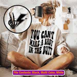 May include: A white sweatshirt with the text "YOU CAN'T MAKE A BABY IN THE BUTT" in black. A black and white circular graphic with a skeleton hand making a peace sign and a lightning bolt is visible. A black mug is held.