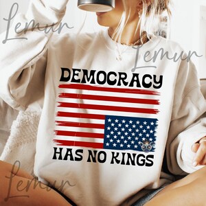 May include: A white sweatshirt with a distressed American flag design and the text "DEMOCRACY HAS NO KINGS".