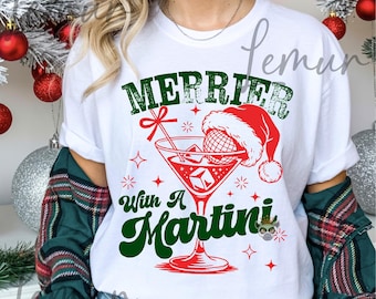 May include: White t-shirt with a festive design. The graphic features the words "Merrier With A Martini" in green and red, along with a martini glass wearing a Santa hat. The design includes stars and a bow.