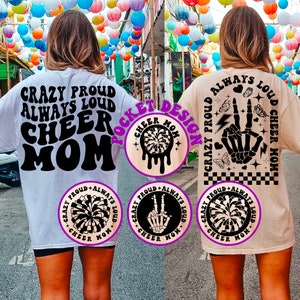 May include: Two white and beige t-shirts with black text and graphics. The front of the white t-shirt says "Crazy Proud Always Loud Cheer Mom". The back of the beige t-shirt says "Crazy Proud Always Loud Cheer Mom" with a skeleton hand making a peace sign and a lightning bolt. The pocket design on the white t-shirt is a black and white graphic of a cheerleading pom-pom.