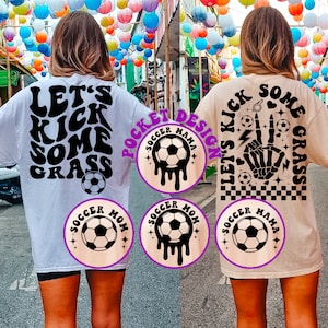 May include: Two white t-shirts with black text and graphics. The front of the shirt says "Soccer Mom" with a soccer ball graphic. The back of the shirt says "Let's Kick Some Grass" with a soccer ball graphic. The second shirt has a similar design with the text "Soccer Mama" and "Let's Kick Some Grass" with a skeleton hand graphic.
