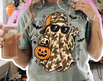 May include: A sage green t-shirt featuring a graphic of a ghost wearing a camouflage baseball cap and black sunglasses. The ghost holds an orange jack-o'-lantern bucket and is surrounded by black spiderwebs, bats, and a small lemur illustration.