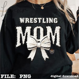 May include: Black sweatshirt with the words "WRESTLING MOM" in white, textured letters outlined in black. A large white bow with black trim is centered below the text. A small emblem of a lemur is on the right side. The words "FILE: PNG" are at the bottom.