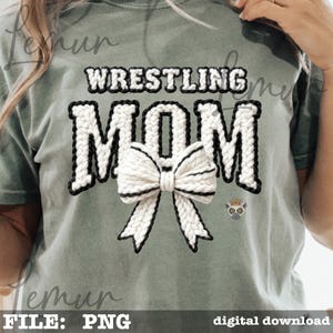 May include: A sage green t-shirt with the words "WRESTLING MOM" in a white, textured font with black trim. A white bow with black trim is centered below the word "MOM". A small lemur graphic is on the lower right.