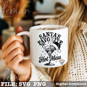 May include: White ceramic mug with the text "SANTA'S FAVORITE HOT MESS" in black. The design includes a martini glass with a Santa hat and stars. The mug is held by a person.