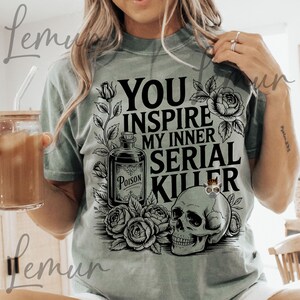 May include: Sage green t-shirt with a graphic design. The design includes a skull, roses, and a poison bottle, with the text "YOU INSPIRE MY INNER SERIAL KILLER". The shirt is a casual wear item.