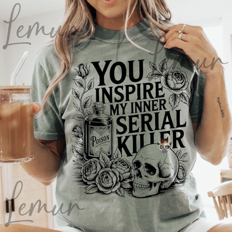 May include: Sage green t-shirt with a graphic design. The design includes a skull, roses, and a poison bottle, with the text "YOU INSPIRE MY INNER SERIAL KILLER". The shirt is a casual wear item.