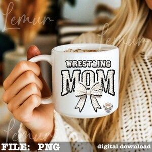 May include: White ceramic mug with the words "WRESTLING MOM" in black and white, featuring a bow graphic. The mug is held by a person.