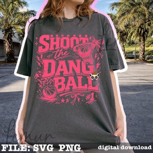 Puede incluir: Camiseta gris oscuro con un dise&ntilde;o rosa con tem&aacute;tica de baloncesto. El dise&ntilde;o incluye el texto "Shoot the Dang Ball", una canasta de baloncesto, una pelota de baloncesto y el n&uacute;mero "#1".