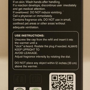 May include: Black box with white text detailing first aid, use instructions, and safety warnings for fragrance oils. Includes a QR code labeled "SCAN ME" and two product certification logos. The box warns against placing objects within 12 inches of the warmer.