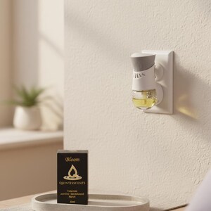 May include: An air freshener plugged into a wall outlet, with a gray and white design and a clear container filled with yellow liquid. A black box with gold text and the word "Bloom" is on a gray tray. The box also says "QUINTESSCENTS".