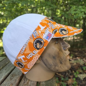 May include: A white cap with an orange brim featuring a repeating pattern of the Tennessee Volunteers logo, the word VOLS, and checkered accents. A small white tag on the side displays the text MADE IN U.S.A. with a small American flag graphic.