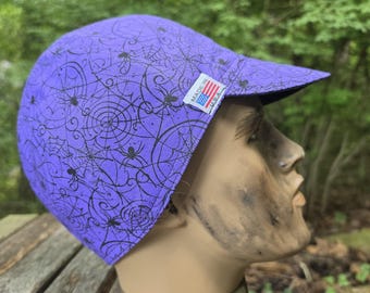 May include: A purple baseball cap featuring a black spiderweb and spider pattern. The cap has a stiff brim and a small white rectangular tag on the side with a graphic of the American flag and the text MADE IN U.S.A. printed in black ink.