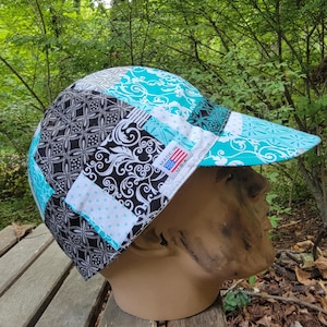 Può includere: Un cappellino da baseball patchwork con un design turchese, nero, bianco e grigio. Il cappellino presenta una varietà di motivi, tra cui forme floreali e geometriche. La visiera è turchese con un motivo floreale bianco. L'etichetta recita "MADE IN USA."