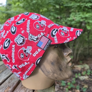 May include: A red welding cap featuring a repeating pattern of University of Georgia Bulldogs logos, football helmets, and the text UGA and GEORGIA. A small rectangular tag on the side displays the American flag and the text MADE IN USA.