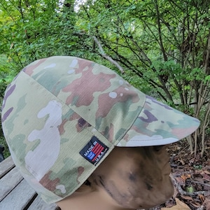 May include: A camouflage pattern flat cap with a short brim. The cap features a mix of green, brown, and tan colors. A small tag on the side reads "MADE IN USA" with an American flag design.