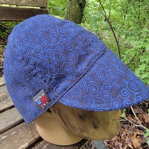 May include: A blue cap with a swirling pattern. The cap is made of fabric and has a small tag that says "Made in USA". The cap is sitting on a wooden surface.