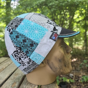 May include: A cycling cap featuring a patchwork design of black, white, and bright blue fabric panels with floral, geometric, and polka dot patterns. The cap has a short brim with blue stitching and a small black tag reading MADE IN U.S.A.