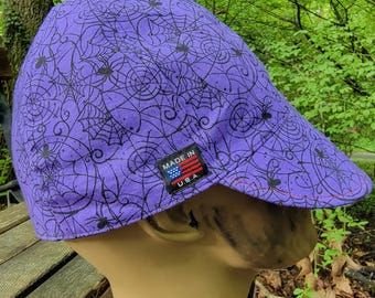 May include: A purple cap with a black spiderweb and spider design. The cap has a small tag that reads "MADE IN U.S.A." The cap is on a mannequin head.