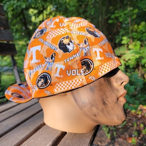 Puede incluir: Gorra de béisbol naranja y blanca con el logotipo de los Tennessee Volunteers, un gráfico de perro sabueso y la palabra "VOLUNTEERS". La gorra tiene un patrón de cuadros blancos y negros en el borde y se muestra en una cabeza de maniquí.