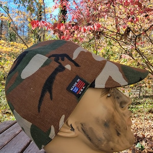 May include: Camouflage pattern baseball cap with a curved brim. The cap features a brown, green, and tan color scheme. A small tag on the side reads "MADE IN U.S.A." in white text with an American flag graphic.