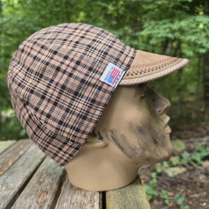 May include: A cycling cap featuring a tan and black plaid pattern with a tan brim. The brim is accented with decorative red and black stitching along the edge. A small white tag on the side displays the text Made in U.S.A. with a flag graphic.