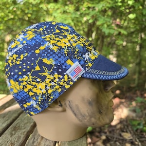 May include: A navy blue and yellow patterned cycling cap featuring the University of Michigan block M logo and a splatter design. The cap has a short, dark blue brim with light blue stitching and a small white tag reading MADE IN U.S.A.
