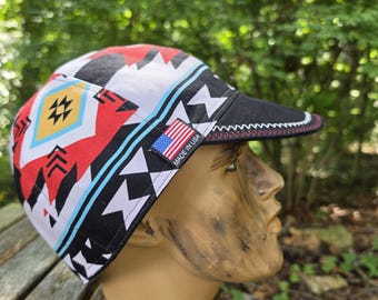 May include: A cycling cap featuring a geometric pattern in red, black, white, and yellow. The cap has a black brim with red stitching and a small rectangular tag on the side displaying the American flag and the text MADE IN USA.