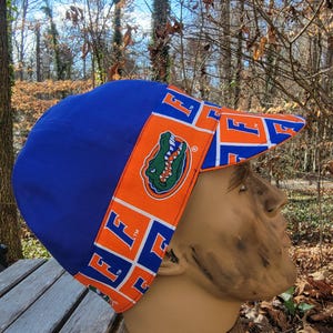 May include: A blue baseball cap with an orange brim and a matching band. The brim and band feature the letter "F" in white and the image of a green alligator. The cap is displayed on a mannequin head.