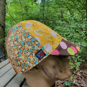 May include: A patchwork newsboy cap with a variety of floral and geometric patterns in shades of yellow, brown, and blue. The brim features brown and pink polka dots. A small tag reads "MADE IN U.S.A."