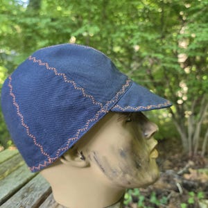 May include: A navy blue cycling cap featuring a short, stiff brim and decorative orange zigzag stitching along the seams. The cap is constructed from a durable fabric and is displayed on a mannequin head to show its rounded, fitted shape.