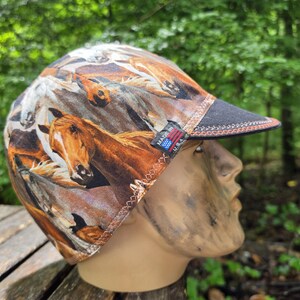 May include: A welding cap featuring a horse print in shades of brown, tan, and white. The cap has a black brim with orange stitching and a small "Made in USA" tag. The cap is displayed on a mannequin head.